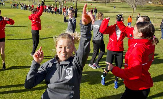 A group of Hamline cross country athletes doing warm-up exercises on a large green lawn