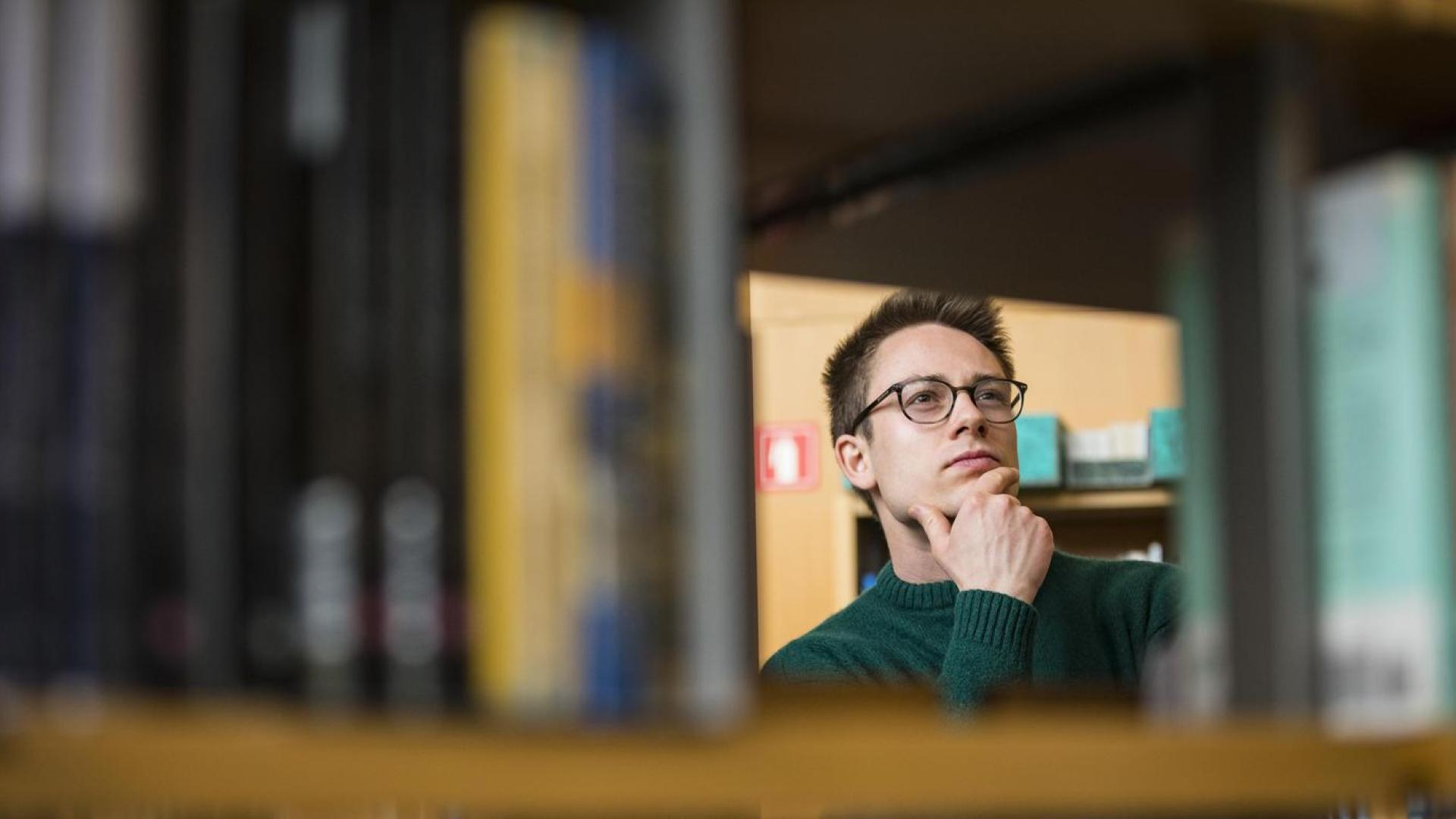 An individual wearing glasses and peering at a library shelf with their hand on their chin